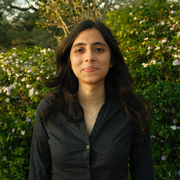 Young woman with dark hair in a patterned shirt against green foliage with small flowers.