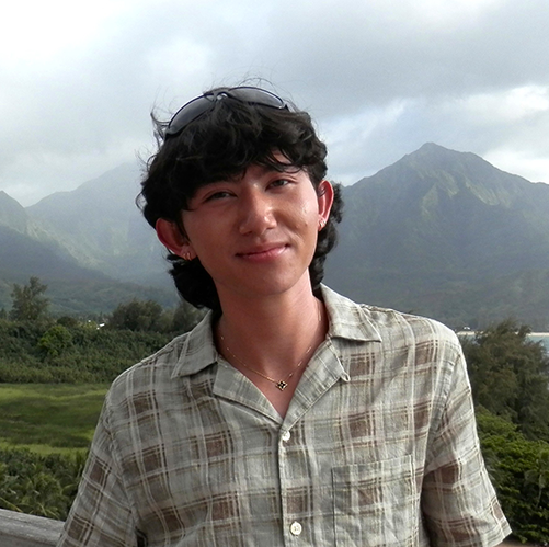 Smiling young man in plaid shirt against a backdrop of lush green mountains.