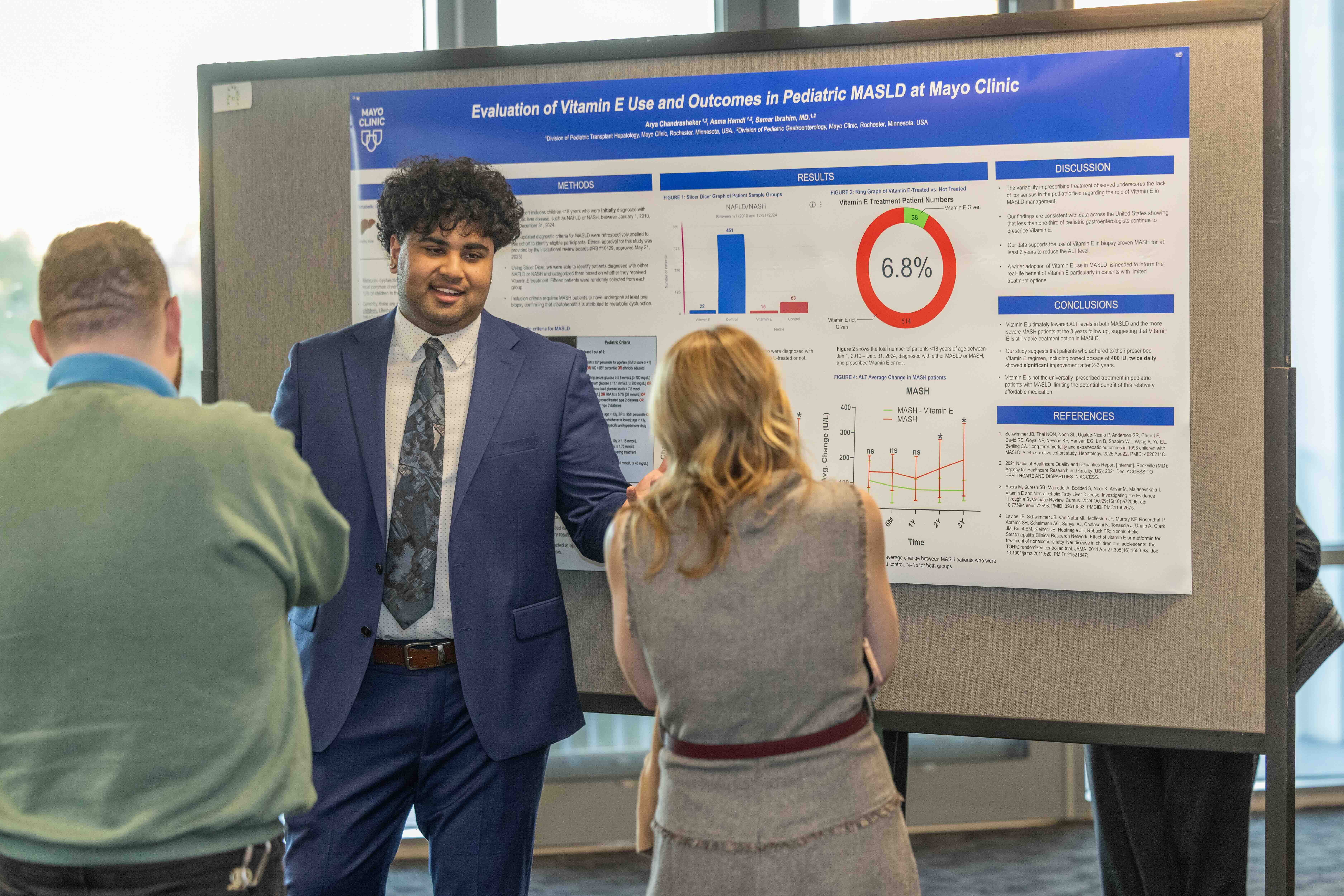 Smiling man in a suit presents a research poster to two people, one seen from behind.