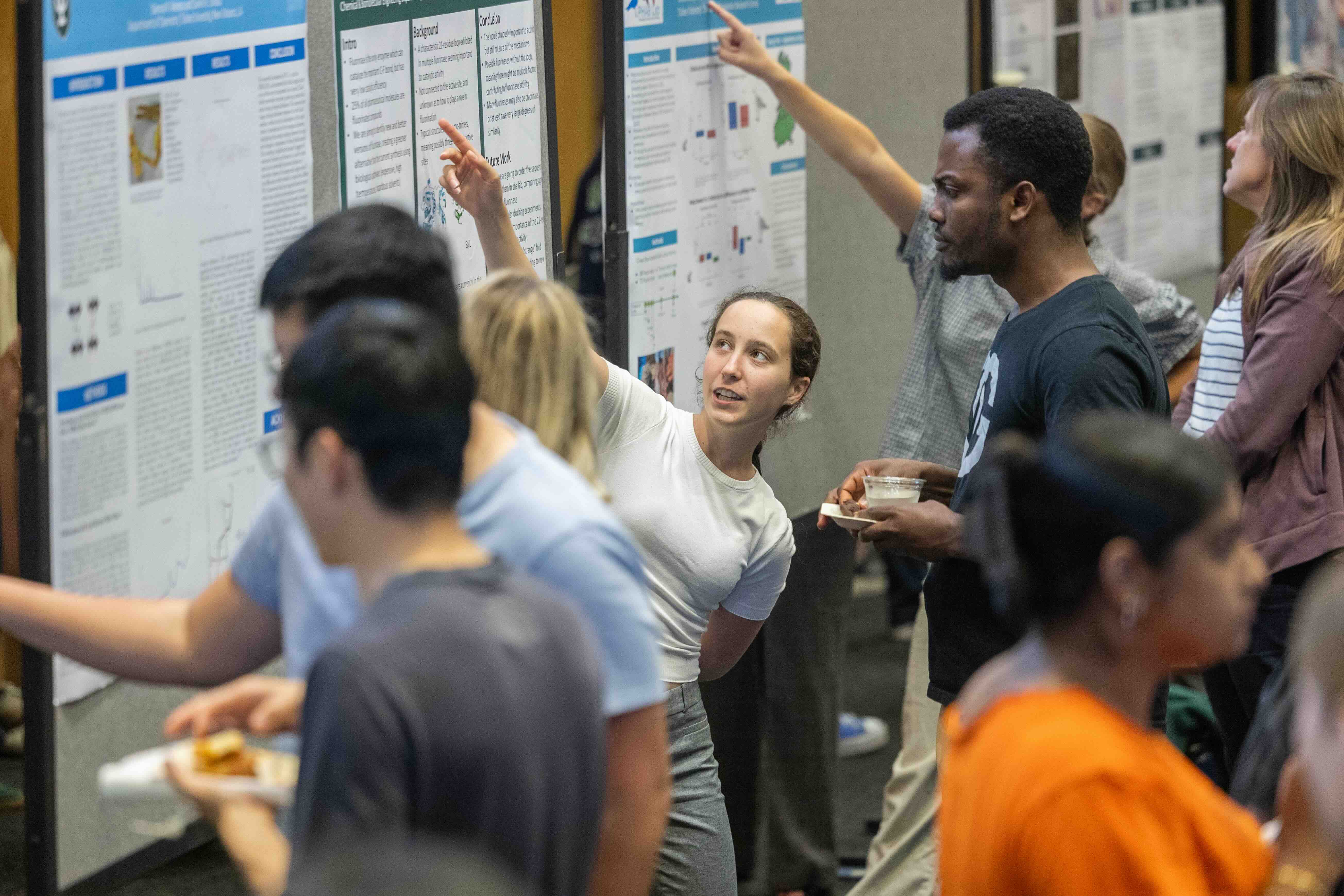 Diverse group of students and professionals at a poster session, many pointing at displays.