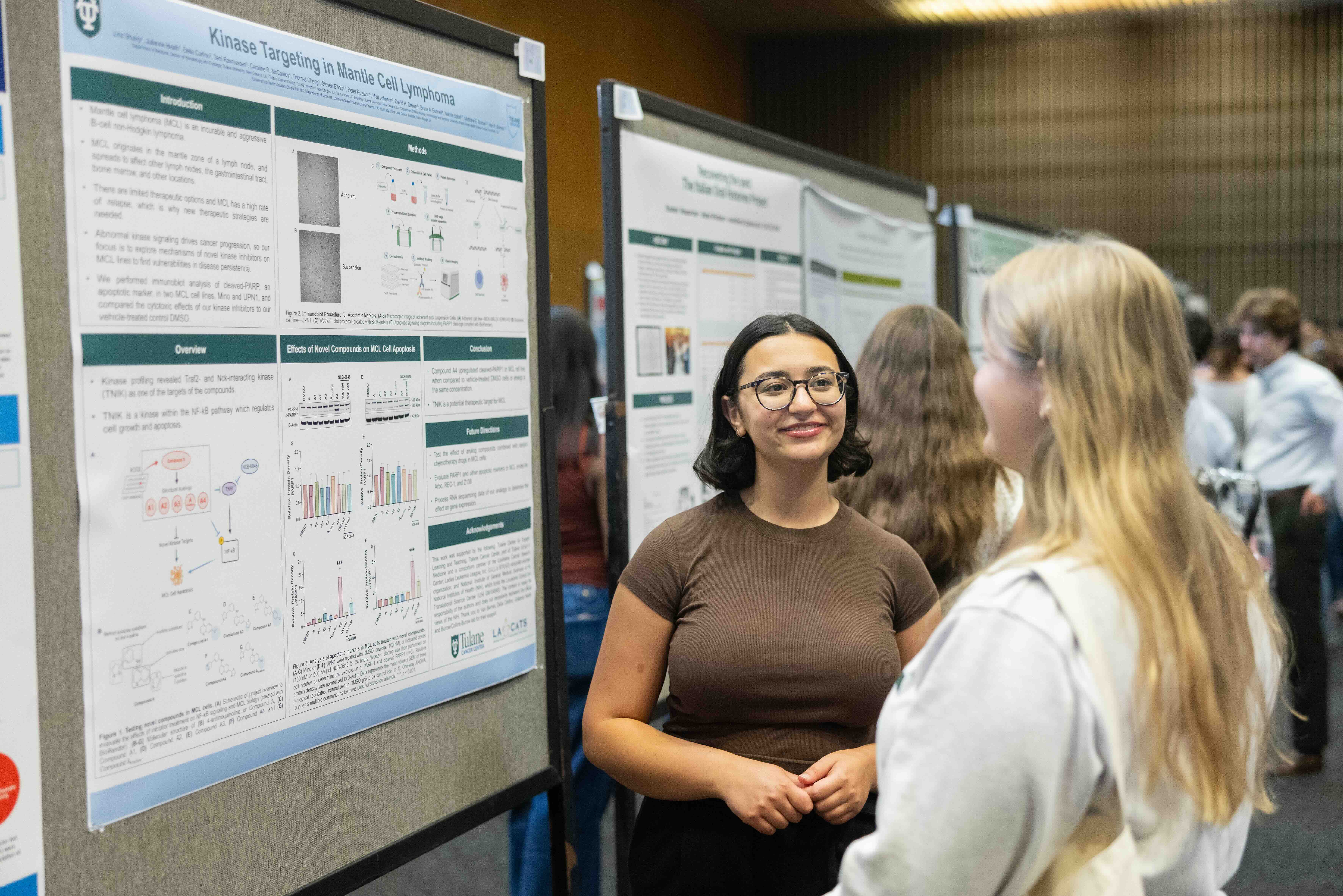 Two young women talking at a scientific poster session, one smiling next to her research poster.